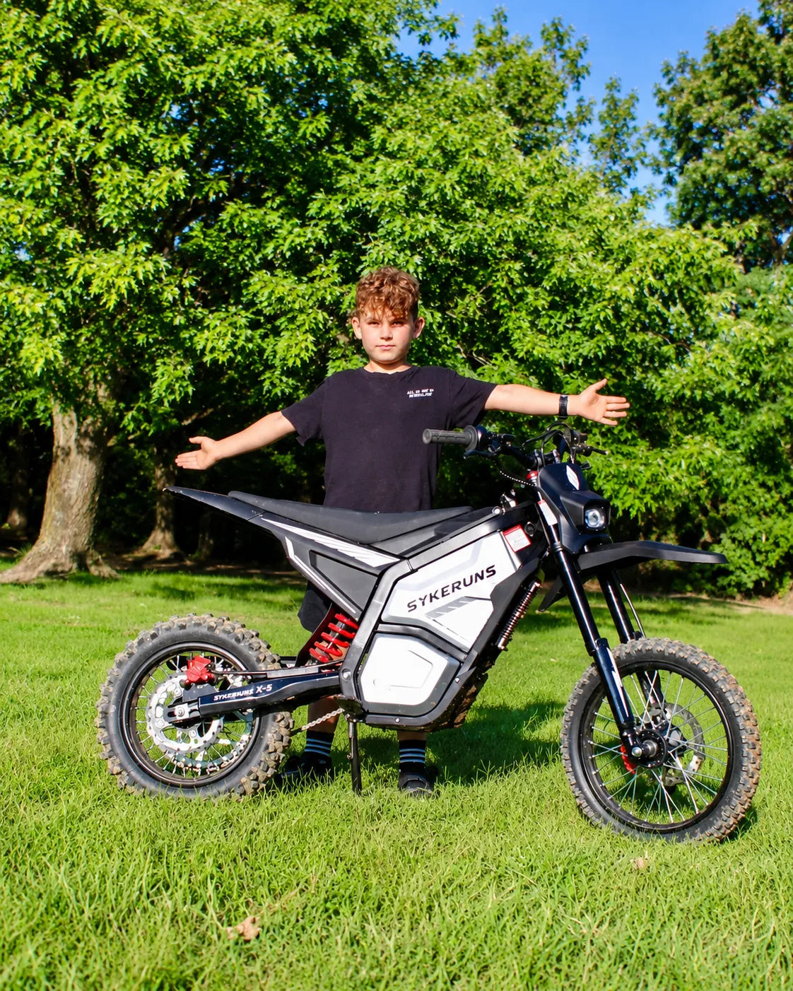 Young rider standing with a SYKERUNS X5 electric dirt bike on a grassy field