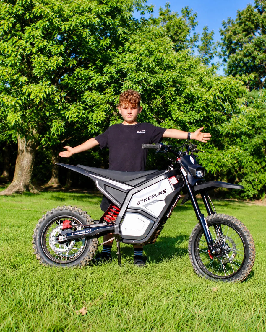 Young rider standing with a SYKERUNS X5 electric dirt bike on a grassy field